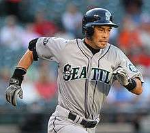 A man in a gray baseball uniform running to the right. The uniform reads "Seattle" across the front, and has a sleeve patch with a teal and silver compass surrounded by "Seattle Mariners". The man is wearing a dark batting helmet.