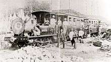 A black-and-white photo of a locomotive carrying and flanked by workers.