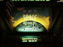 A photograph of a close-up of an arcade screen. A video monitor rests at the bottom of the image, and the monitor's displayed graphics are reflected onto artwork of a grey moon surrounded by yellow and green colors. The reflected graphics display the phrase "Space Invaders".