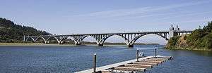 A large river flows under a long elegant bridge with multiple arches. A dock and boat are in the foreground.