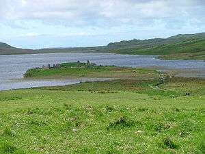 A small island in a lake lies offshore from green fields. A small wooden footbridge leads to the islet which contains various stone ruins including at least two gable ends.