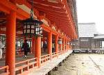 A wooden corridor on stilts with red beams and a red handrail. There are metal lanterns hanging from the ceiling.