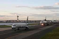 Five jumbo airplanes wait in a line on a runway next to a small body of water. Behind them in the distance is the airport and control tower.