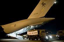 Colour photo of a truck with a sign marked with Japanese characters over its cargo compartment driving away from the rear ramp of a grey aircraft at night.