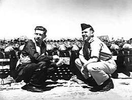 Two men crouching opposite each other, one in flying gear and the other in light-coloured uniform with forage cap