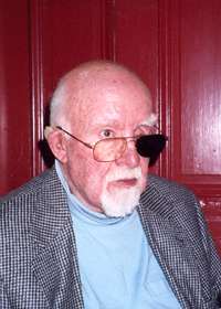 Head and shoulders of elderly man wearing glasses with one black lens. The man is white-haired and mostly bald, and sports a goatee and mustache.