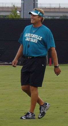 Color full-length photograph of stocky, well-tanned white man (Jack Del Rio), wearing teal blue Jacksonville Jaguars teal t-shirt, black Bermuda shorts and teal and white sun visor, walking across playing field.