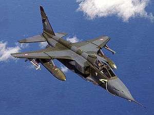 Top forward view of dark-green and blue-grey aircraft overflying sea, above scattered clouds.