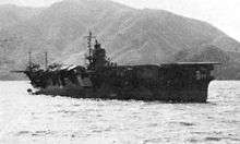 Photograph of a ship floating in the ocean taken from a location on the ocean surface some distance away. The upper deck of the ship is flat except for a superstructure visible near the center of the ship. The shoreline and some mountainous terrain are visible in the distance behind the ship; the sky is visible above the mountains.