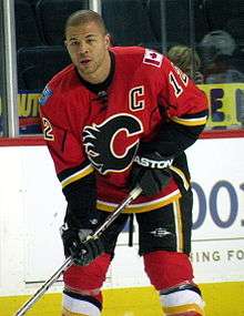 Hockey player in red and black uniform, with a "C" in the middle and no helmet on. He holds his stick diagonally.