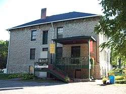 A different side of the same stone building, with stairs and a metal porch to the right from which flowering plants hang. There are banners on the porch identifying it as the Jell-O Museum in Le Roy, New York