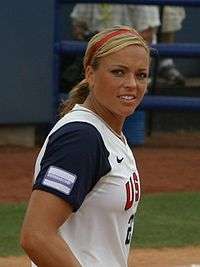 A blond woman wearing a blue and white softball uniform along with a red headband. Dermabond and Nike logos appear on her uniform, as does "US" and part of her number