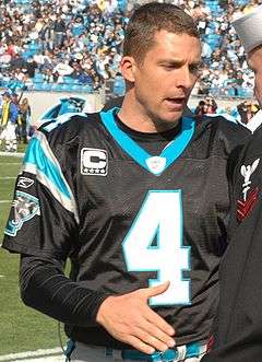 A man, in football uniform but not wearing a helmet, is standing at midfield, preparing to shake hands with another person.
