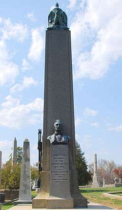 A large obelisk in a graveyard, with a bust of Tyler. The black structure visible behind the left side of Tyler's obelisk encloses the grave of James Monroe.