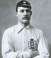 A black-and-white upper-body photograph of a man wearing a white shirt and a dark cap. He has a moustache and his arms are folded.