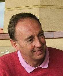 A smiling middle-aged white man with short hair, wearing a pink shirt and red pullover, looking to his left with the tip of his tongue between his lips