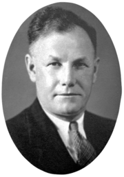 A head and shoulders, black and white photographic portrait of a white man with receding dark hair