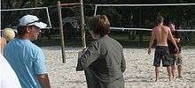 On the left in the foreground, a left facing man in a light blue shirt faces a woman with short brown hair and a green jacket. In the background, a shirtless male teenager with black shorts talks to a shorter man wearing a black shirt. The people are standing on a volleyball court with the net stretched across the background.