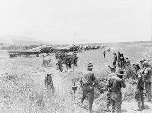 A line of Dakotas on a grass airstrip. Men wearing slouch hats file off a plane. A jeep drives along the strip. Other men in uniform and civilians look on.