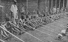 A row of more than a dozen children holding wooden looms stretches into the distance.