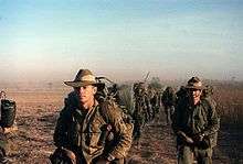 A group of men wearing green military uniforms walking across barren ground.