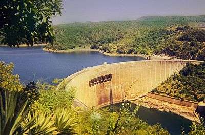 An impressive dam, built in a semi-circular shape, viewed from a vantage point above and to the right of its front. A large lake can be seen on the other side of the dam as well as a river before it.