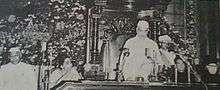 Photo of man in white cap/robe standing to speak at table