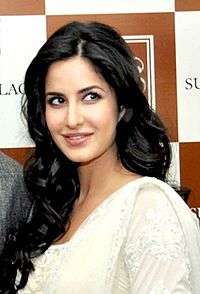 A woman dressed in a white salwar kameez (traditional Indian dress) with her hair down is smiling and looking away from the camera.