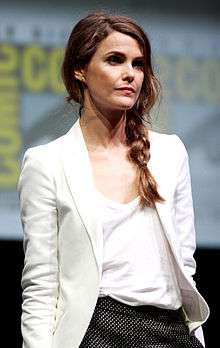 A young woman in a white top looks at the camera. Behind her is a wall with white and yellow logos.