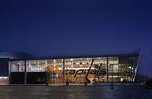 A modern glass building in the shape of a rhombus with the words Washington Capitals large across the front lit up under a dark blue evening sky.