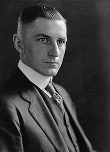 A man in his late twenties with dark, short, slicked-back hair wearing a black coat and tie and white shirt