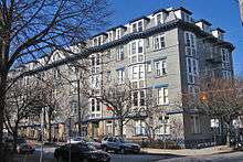 A five-story gray building with blue trim and a mansard roof, seen from across the street and partially obscured by bare trees on the left