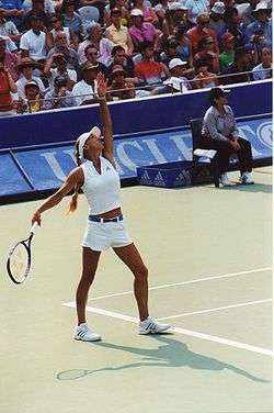 Anna Kournikova playing tennis in white outfit. Left hand is extended as if she has just tossed a ball and right hand is cocking back for the serve.