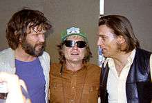 Three men. From left to right, the first man has brown hair and beard. He wears a blue T-shirt and a white jacket and is looking at the man in the middle. The man in the middle wears a green cap and shades, and long red hair. He wears a brown T-shirt. The man at the right has brown hair, he looks at the man at the middle. He wears a white shirt and a black letter jacket.