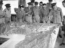 Men in military-style uniforms stand around a terrain model while one man points out terrain features with a pointer