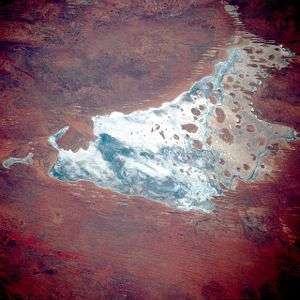 Satellite photograph of an irregularly-shaped body of water, containing numerous small islands, and surrounded by red land marked by numerous parallel lines.