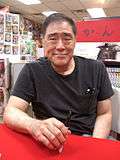 A smiling, middle-aged man with thin dark hair sitting at a red table wearing a black T-shirt.