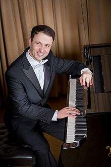 A dark haired man in a tailcoat and a kummerbund, dark bow tie, both hands in the pockets of light-coloured trousers.