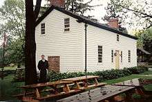 A white house in three-quarter perspective.  In the foreground are several picnic benches.  To the left is a tall tree with a man standing beside it.  To the right, partly obsured by the house, is an old well.