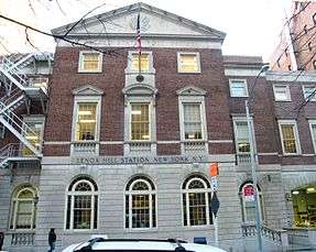 A three-story brick building with grayish-brown stone trim and an American flag hung from the front on a city street. The middle of the front, with a pointy top and round-topped windows, sticks out slightly. "UNITED STATES POST OFFICE" is carved into the stone near the top, and "Lenox Hill Station New York N.Y." near the street. There is a pale blue fire escape on the left.