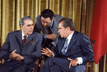 Two older men in suits sit next to each other, while a third stands behind leaning in to listen to the right man talk.