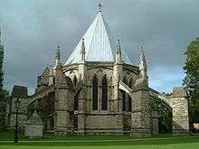  An external view of the Chapter House at Lincoln against a dark cloudy sky. The polygonal building has a steeply pointed lead roof, paired pointed windows and huge flying buttresses with splay out all around the building like spider's legs.