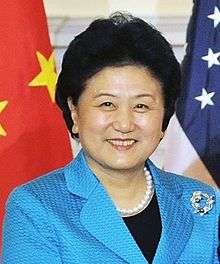 a smiling, aged woman woman, wearing a blue dress (and a black shirt under it), white necklace and standing in front of the American and Chinese national flags