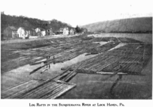 Several rafts made of long logs lashed together are bunched together near the bank of a large river. A row of houses lines the river bank, at the bottom of hill paralleling the river. Two men are standing on one of the rafts.