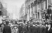 Black and white photograph of a crowded street, taken at eye-level. Clothes can be seen hanging at stalls along the sides of the street.