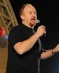 A balding man with red hair and a red goatee, wearing a black T-shirt, speaks into a microphone.