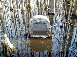 A granite marker sits in the swamp water surrounded by trees with an inscription detailing its use marking the point of beginning of the Louisiana Purchase surveys and its placement in 1926