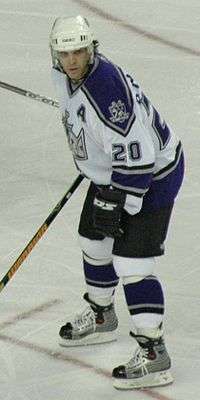 Hockey player in white and blue uniform. He stands shoulder-width apart and stands in a readied stance.