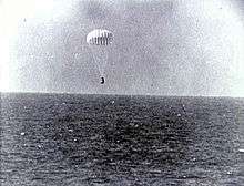 A conical black spacecraft falling towards the surface of the ocean under a single white parachute, seen from some distance away. Very little detail can be seen.