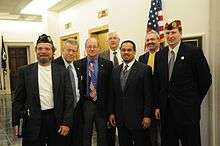 Man in his forties wearing a dark suit, white shirt, striped tie and gray vest. He has a small beard or goatee and is standing with six men, all wearing suits and two wearing small hats from the National Guard and the letters "D. A. V."